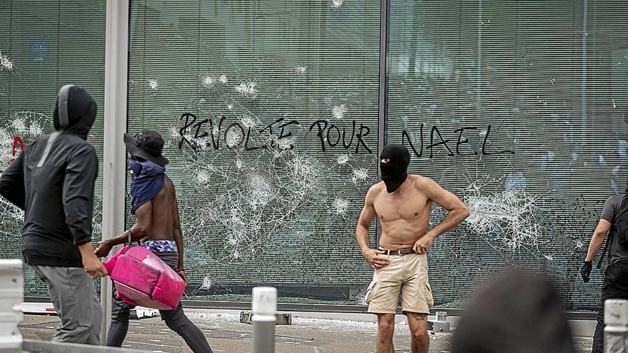Un grupo de encapuchados delante de un escaparate con una pintada que dice: ‘Revuelta por Nahel’.