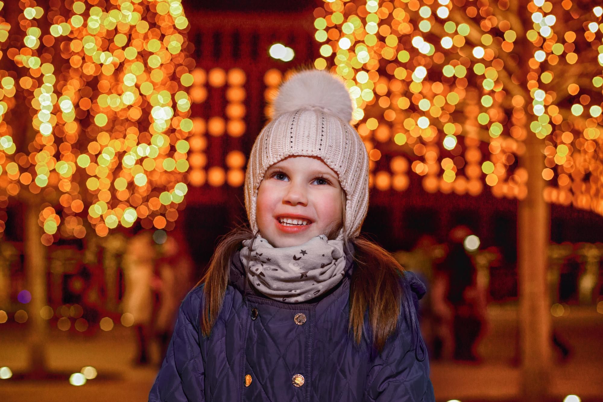 Una niña protege sus oídos del frío con un gorro en una calle con luces de Navidad.