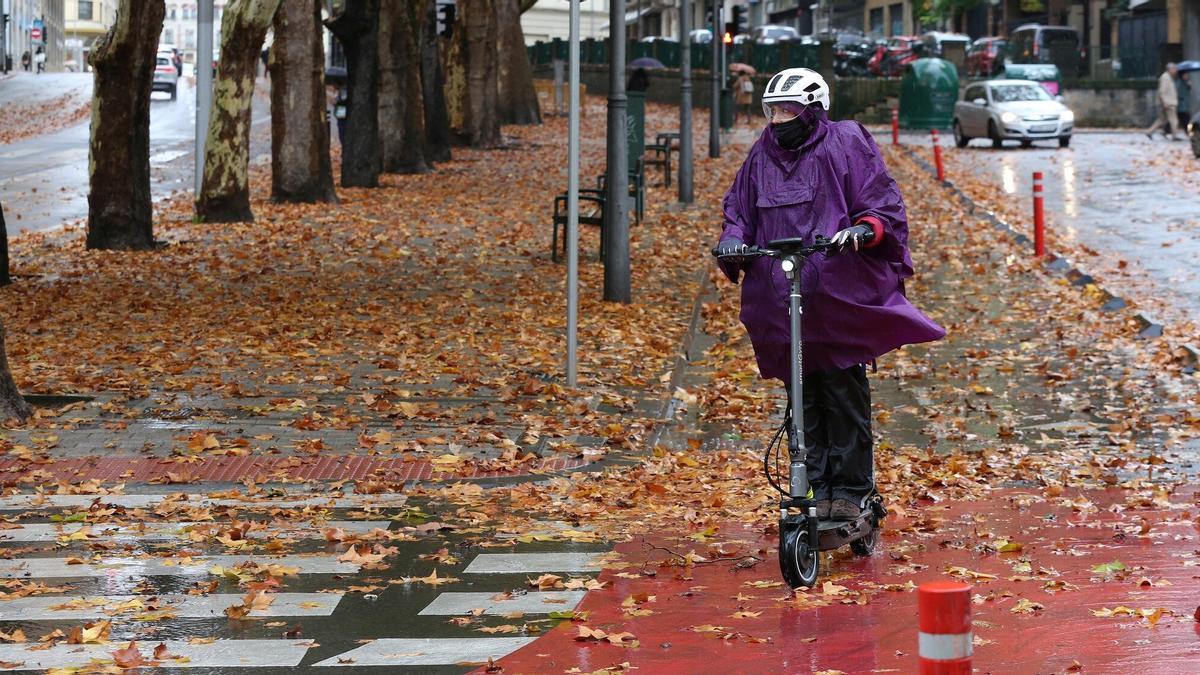 Una mujer en patinete se protege de la lluvia con un chubasquero.