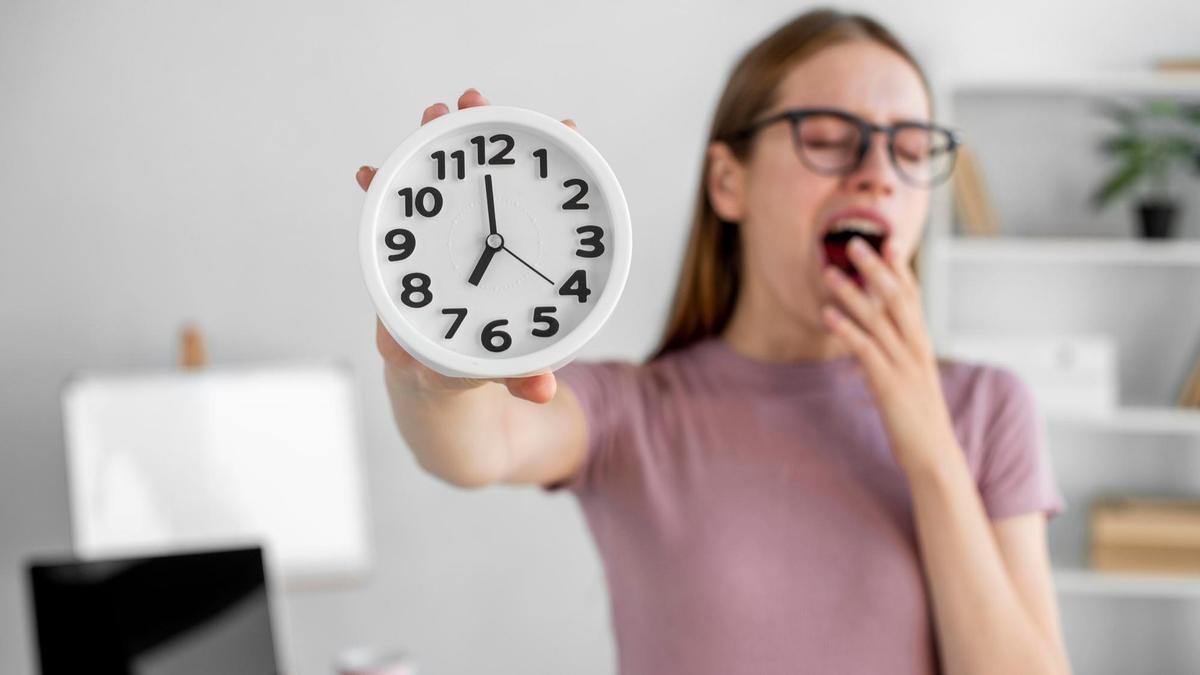 Una mujer bosteza mientras sujeta un pequeño reloj.