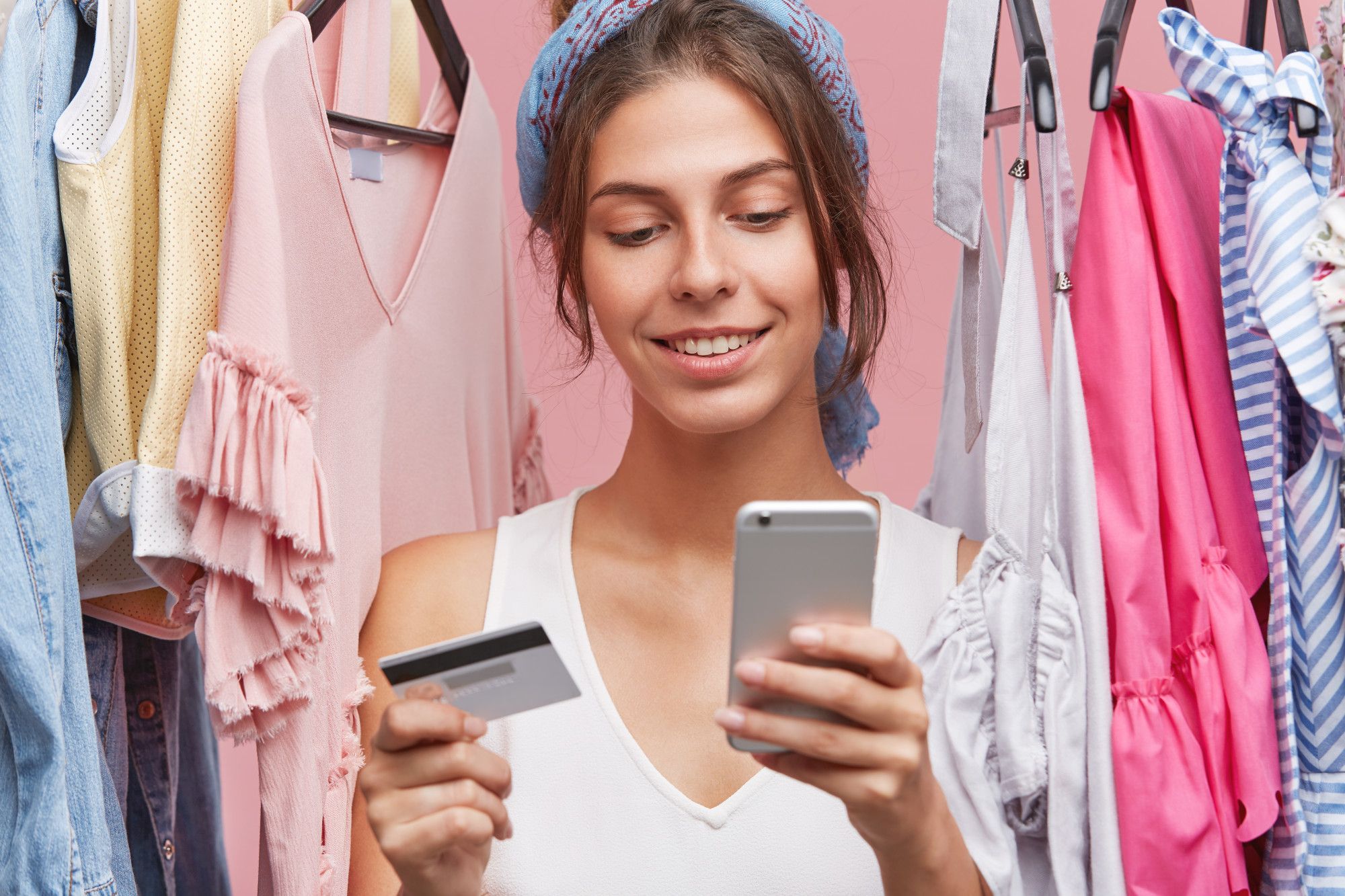 Una joven hace una compra online junto a un perchero lleno de ropa.